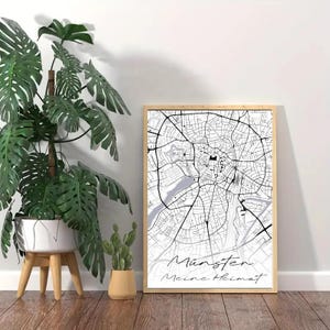 Puede incluir: Una impresión de mapa de ciudad en blanco y negro con el texto "Münster Meine Heimat" en un marco de madera. La impresión está colgada en una pared blanca con una planta en maceta delante.