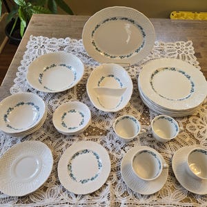 Puede incluir: Un juego completo de vajilla de porcelana blanca con un patrón de tejido de cesta y detalles florales azules. El juego incluye platos, cuencos, tazas, platillos y fuentes para una experiencia gastronómica formal.