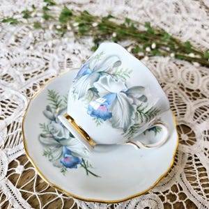 May include: A white teacup and saucer with a floral design featuring blue and white orchids. The cup and saucer have a gold trim.