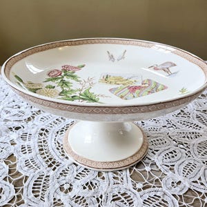 May include: A white ceramic cake stand with a brown and white patterned rim. The stand features a floral design and a bird illustration.