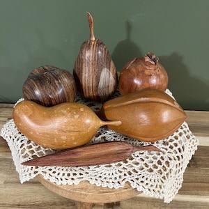 May include: A collection of six hand-carved wooden decorative fruit sculptures, including a pear, a pomegranate, and various rounded and elongated shapes, featuring natural wood grain patterns in shades of brown, tan, and dark mahogany.