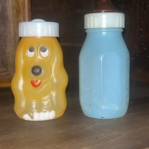May include: A yellow plastic container shaped like a dog with a white screw-top lid and a light blue plastic baby bottle with a white collar and a brown nipple, featuring embossed measurement markings on the side indicating up to 4 ounces.