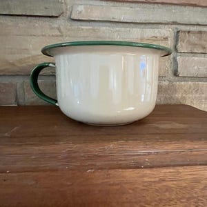 Può includere: Un vaso da notte in smalto color crema caratterizzato da un bacino ampio e arrotondato e da un manico curvo. Il bordo del bacino e il manico sono rifiniti con un bordo verde scuro, creando un netto contrasto con il corpo chiaro.