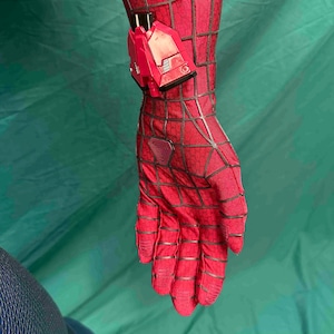 May include: A red fabric glove featuring a black web pattern and a triangular emblem on the back of the hand. A rigid, bright red mechanical device with black accents is attached to the wrist area of the glove, extending over the forearm.