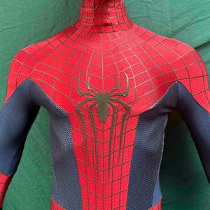 May include: A full-body spandex suit featuring a vibrant red chest and shoulder section with a black web pattern and a large black spider emblem in the center. The sides and arms are a textured dark blue material with a honeycomb-like pattern.