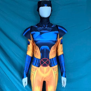 Puede incluir: Un disfraz de cuerpo entero en amarillo, azul y negro, con un tocado y un body con un emblema circular. El disfraz tiene mangas largas y se muestra en un maniquí. El diseño incluye una combinación de colores vibrantes y un tema de superhéroe.