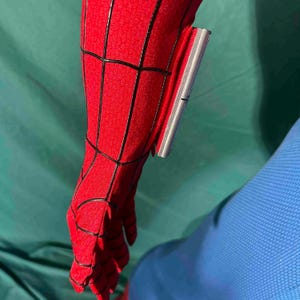 Puede incluir: Primer plano de una manga de disfraz de Spider-Man rojo y negro. La tela roja tiene un patr&oacute;n de telara&ntilde;a con l&iacute;neas negras. Un dispositivo plateado est&aacute; adherido a la manga.