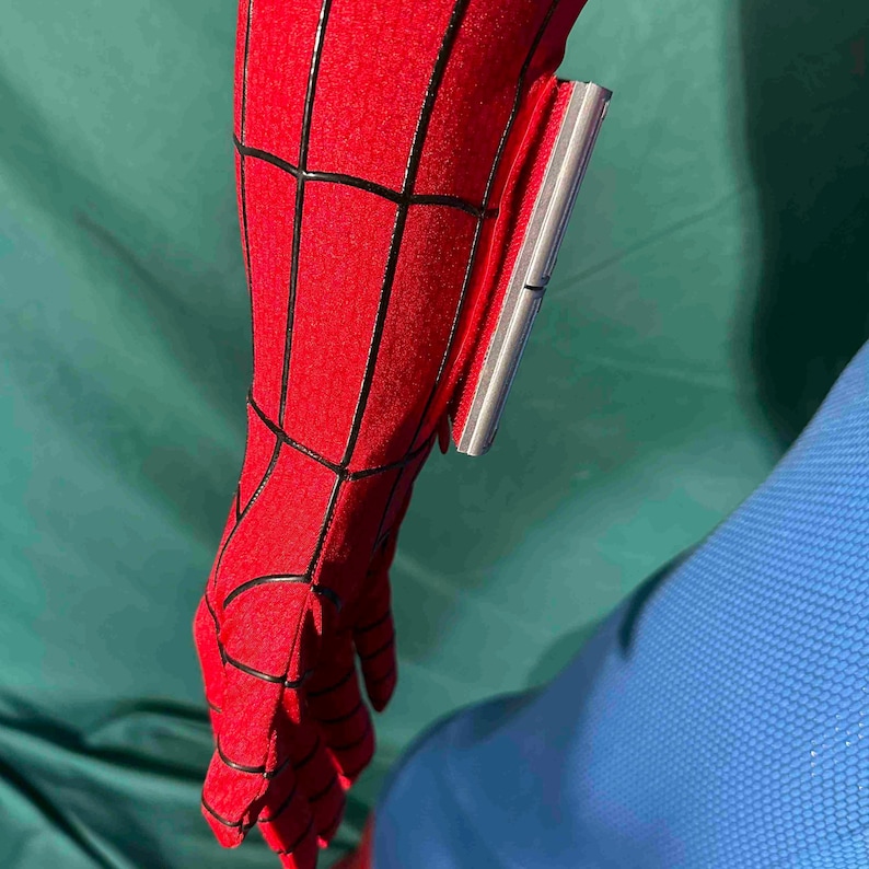 Puede incluir: Primer plano de una manga de disfraz de Spider-Man rojo y negro. La tela roja tiene un patr&oacute;n de telara&ntilde;a con l&iacute;neas negras. Un dispositivo plateado est&aacute; adherido a la manga.