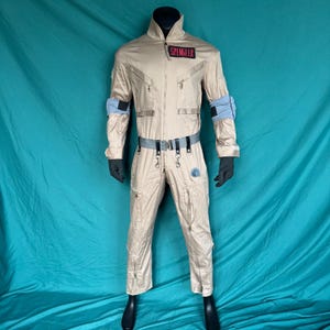 May include: Tan jumpsuit costume with black gloves and boots. The costume features a high collar, multiple pockets, and a gray belt. The name "SPENGLER" is displayed on a red patch. Blue armbands and a circular emblem are also visible.