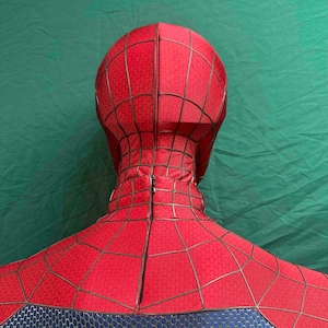 May include: A red full-head mask and upper torso costume piece featuring a black web pattern printed across the fabric. The mask includes a vertical zipper closure running down the back of the neck, with a blue textured section at the bottom.