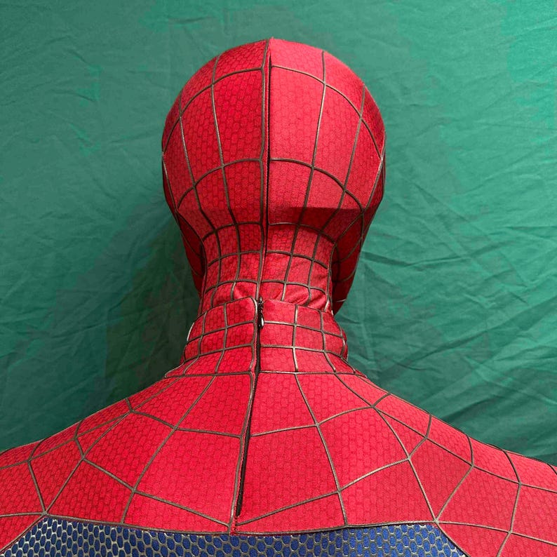 May include: A red full-head mask and upper torso costume piece featuring a black web pattern printed across the fabric. The mask includes a vertical zipper closure running down the back of the neck, with a blue textured section at the bottom.