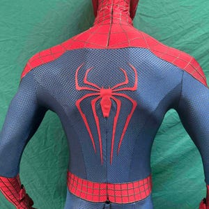 May include: A full-body spandex costume featuring a textured blue base with red sections on the shoulders and waist. A large, elongated red spider emblem is centered on the back, complemented by a black web pattern printed over the red areas.