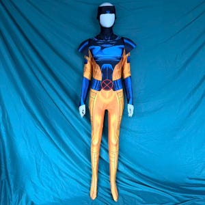 May include: A full-body costume in yellow, blue, and black, displayed on a mannequin. The costume features a black head covering, blue shoulder pads, and a red and black circular emblem on the chest. The legs and arms are yellow with blue accents.