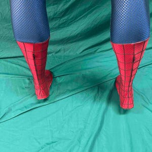 May include: A pair of costume leggings featuring a blue upper section with a textured honeycomb pattern and a red lower section with a black webbed grid design. The red portions extend from the mid-calf down to the feet and include a vertical zipper.