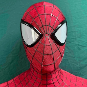 May include: A red full-head mask featuring a black web pattern across the fabric surface. The mask includes two large, almond-shaped eye lenses with a white mesh texture, each framed by a thick, raised black border that defines the facial structure.