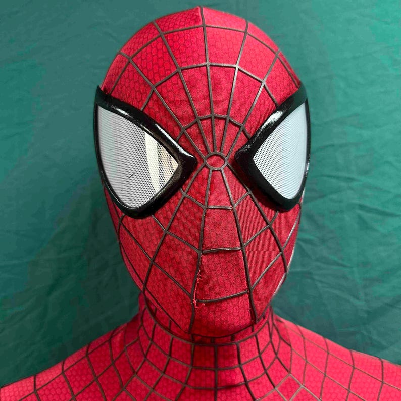 May include: A red full-head mask featuring a black web pattern across the fabric surface. The mask includes two large, almond-shaped eye lenses with a white mesh texture, each framed by a thick, raised black border that defines the facial structure.