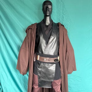 Puede incluir: Un traje de cuero negro con una túnica marrón. El traje presenta un chaleco de cuero negro con cuello en V y un cinturón de cuero marrón con una hebilla plateada.