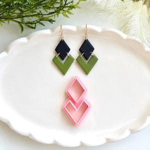 May include: A pair of geometric dangle earrings with black and olive green diamond shapes, hanging from gold-colored hooks. A pink diamond-shaped earring cutter is also present, suggesting a crafting theme. The earrings and cutter are displayed on a white, scalloped plate.