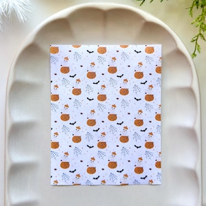 Può includere: Una carta rettangolare con un motivo ripetuto su sfondo bianco. Il design include piccole zucche arancioni, fantasmi bianchi con cappelli arancioni, ragnatele nere e pipistrelli neri sparsi sulla superficie in una disposizione a griglia.