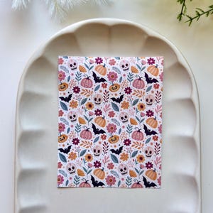 Può includere: Una carta rettangolare con un motivo ripetuto di teschi bianchi, zucche arancioni e rosa, pipistrelli neri e vari fiori colorati nelle tonalità del giallo, rosa e bordeaux su sfondo bianco, posizionata su un vassoio bianco dal bordo smerlato.