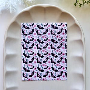 Könnte beinhalten: Ein rechteckiges Blatt Papier mit einem sich wiederholenden Muster aus schwarzen Fledermäusen mit rosa Gesichtern und ausgebreiteten Flügeln, durchsetzt mit kleinen rosa Blüten und zarten blau-grünen Blattzweigen auf weißem Grund.