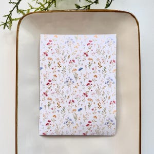 Puede incluir: Una tarjeta rectangular con un fondo blanco cubierto por un patrón floral de flores silvestres rojas, rosas, azules y amarillas. La tarjeta está centrada en un plato color crema con un borde marrón, con vegetación en la esquina superior izquierda.