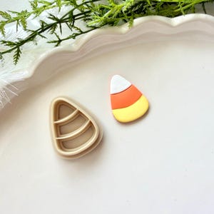 Może przedstawiać: Beżowa plastikowa foremka do ciastek w kształcie cukierka kukurydzianego obok małego, gładkiego elementu w kształcie cukierka kukurydzianego z poziomymi pasami w kolorze białym, pomarańczowym i żółtym na jasnym, jednolitym tle.