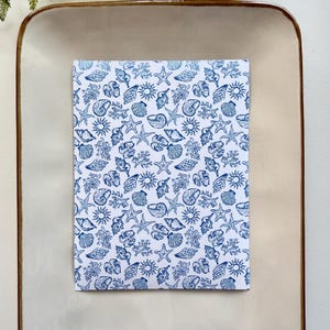 May include: A notebook with a white background and a blue seashell pattern. The pattern includes starfish, shells, coral, and suns. The notebook is rectangular and sits on a light brown tray.