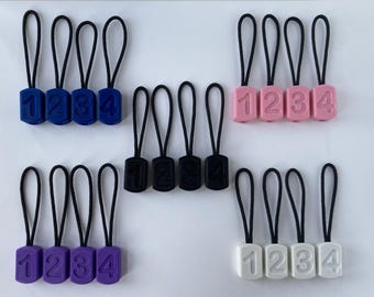 Puede incluir: Cuatro juegos de cuatro tiradores de cremallera con los números 1, 2, 3 y 4. Los juegos son en azul, rosa, negro y blanco.