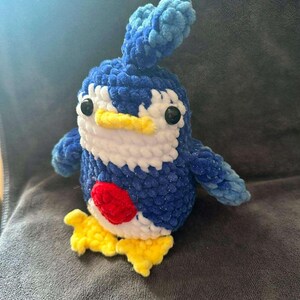 May include: A handmade crochet plush toy shaped like a penguin with dark blue, light blue, and white yarn. It features a yellow beak and feet, black plastic safety eyes, and a prominent red circular patch on its chest. The texture is soft and fuzzy.