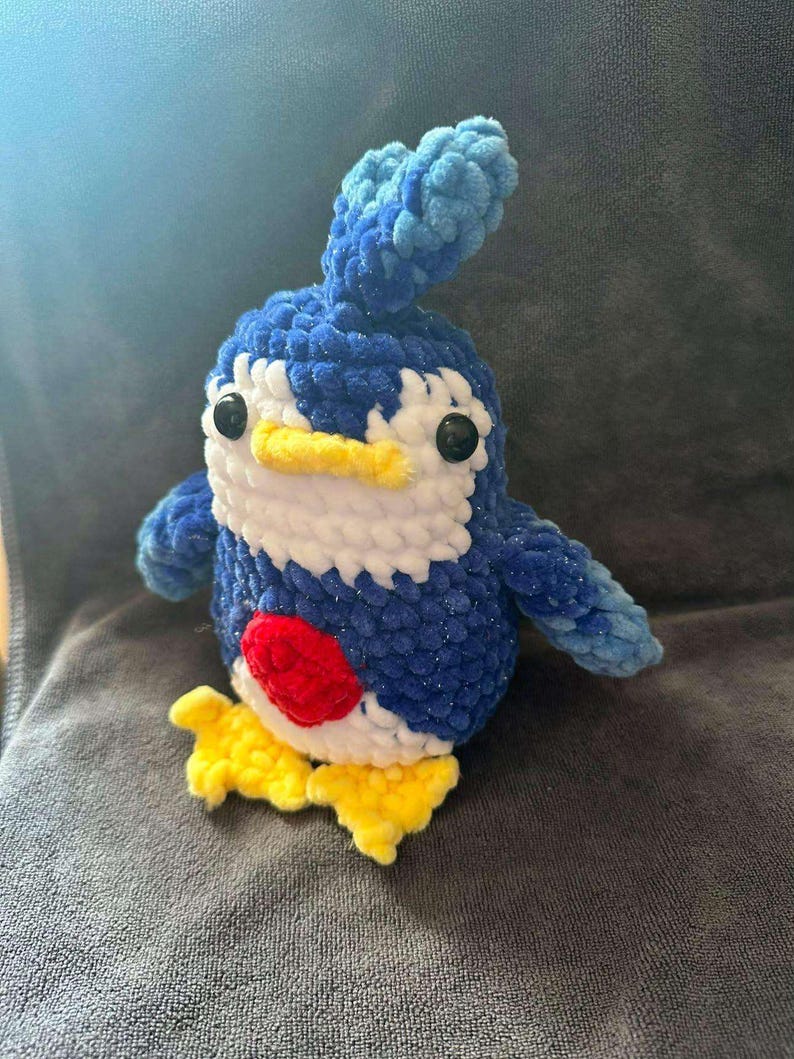 May include: A handmade crochet plush toy shaped like a penguin with dark blue, light blue, and white yarn. It features a yellow beak and feet, black plastic safety eyes, and a prominent red circular patch on its chest. The texture is soft and fuzzy.
