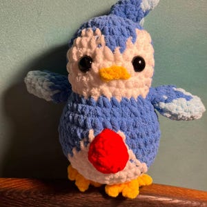 May include: A handmade crochet plush toy shaped like a bird with blue and white yarn, featuring a bright red circular patch on its chest, a small yellow beak, yellow feet, and two black plastic safety eyes. The texture is soft and fuzzy.