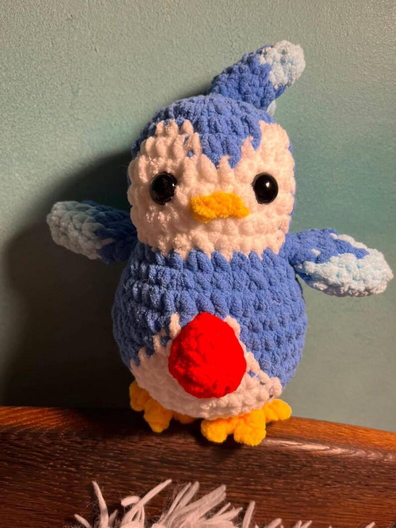 May include: A handmade crochet plush toy shaped like a bird with blue and white yarn, featuring a bright red circular patch on its chest, a small yellow beak, yellow feet, and two black plastic safety eyes. The texture is soft and fuzzy.
