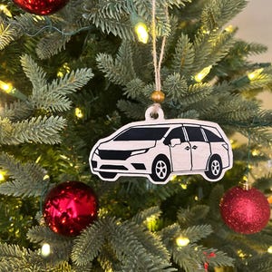 May include: White wooden Christmas ornament shaped like a minivan. The ornament is hanging on a green Christmas tree with red ornaments and white lights.