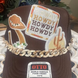 Könnte beinhalten: Braune Trucker-Mütze mit aufgestickten Aufnähern, darunter ein Cowboyhut, ein Kaktus und das Wort "HOWDY". Die Mütze hat ein Kettendetail und ein Etikett "World Famous Foam Front". OSFM-Größe.