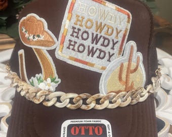 Może przedstawiać: Brązowa czapka typu trucker z haftowanymi naszywkami, w tym kapeluszem kowbojskim, kaktusem i napisem "HOWDY". Czapka posiada łańcuszek i etykietę "World Famous Foam Front". Rozmiar OSFM.
