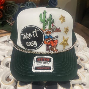 Könnte beinhalten: Grün-weiße Trucker-Mütze mit einer Perlenkette und einer Grafik eines Gitarre spielenden Cowboys, Kakteen und goldenen Sternen. Die Mütze trägt den Text "Take it easy" und "OTTO TRUCKER" auf der Vorderseite.