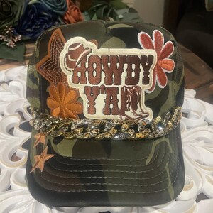 Könnte beinhalten: Camouflage-Trucker-Cap mit den Worten "HOWDY YA'LL" in Braun und Weiß. Die Kappe hat eine Kette über dem Rand und Stern- und Blumenverzierungen. Die Kappe hat eine Netzrückseite.
