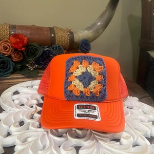May include: Bright orange trucker hat with a crocheted square design in the center. The square features a blue, orange, and cream-colored pattern. The hat has a mesh back and a snap closure. The label reads "OTTO World Famous Foam Front".