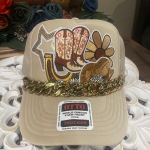 Könnte beinhalten: Beige Trucker-Cap mit goldener Kette, bestickt mit Aufnähern von Cowboy-Stiefeln, einem Hufeisen, einem Stern und einer Gänseblume. Die Kappe hat einen gebogenen Schirm und eine Mesh-Rückseite. Das Etikett lautet "OTTO TRUCKER".
