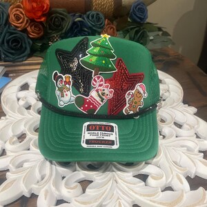 Könnte beinhalten: Eine grüne Trucker-Mütze mit schwarzem Schirm und schwarzem Seildetail. Die Mütze hat weihnachtliche Aufnäher, darunter ein Weihnachtsbaum, Sterne, ein Schneemann und ein Lebkuchenmann. Die Mütze hat einen Schnappverschluss.