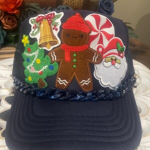 May include: Navy blue trucker hat with Christmas-themed patches. Patches include a gingerbread man, Santa Claus, a Christmas tree, a bell, and a red and white swirled candy. The hat has a blue chain detail along the brim.