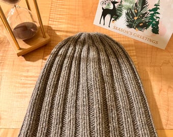 May include: A grey ribbed knit beanie hat positioned below a wooden hourglass and a Season's Greetings card featuring a black silhouette of a reindeer and green pine trees on a light background.