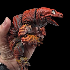Pode incluir: Figura de dinossauro articulada vermelha e laranja detalhada com uma superfície texturizada. A figura tem uma cabeça detalhada com a boca aberta e é segurada contra um fundo preto. Os olhos da figura são brancos com uma pupila preta.