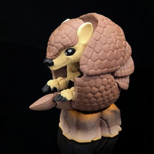 May include: A detailed figurine of an armadillo in a curled position. The figure has a brown, textured shell and a light tan face, ears, and underbelly. The armadillo's claws are black, and it is posed on a rock-like base.