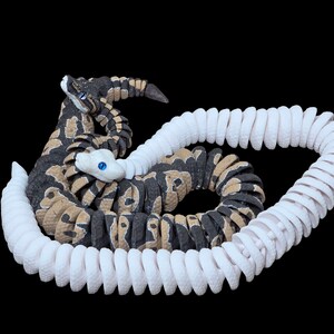 May include: A detailed sculpture of a rattlesnake with a coiled white tail. The snake's body is patterned with black, tan, and brown markings. It has a white head with blue eyes, set against a black background.