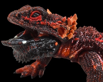Pode incluir: Um close-up de uma figura de lagarto vermelho e preto com uma superfície texturizada e escamosa. O lagarto tem a boca aberta, revelando dentes afiados.