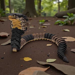 Puede incluir: Una detallada cobra de juguete articulada, con una combinación de colores negro, beige y amarillo. La serpiente está posada en el suelo de un bosque con hojas caídas y vegetación de fondo. La cabeza de la serpiente está levantada y su cuerpo curvado.