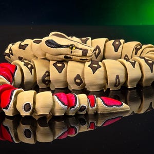 May include: A wooden articulated snake toy with a tan body, dark brown markings, and a red and white patterned tail. The snake's head is tan with dark brown and black details, and yellow eyes. The toy is reflected on a black surface.