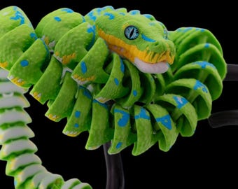 Pode incluir: Uma cobra verde e azul com detalhes amarelos está enrolada em torno de um galho preto. A cobra tem um olho azul brilhante e uma boca branca.